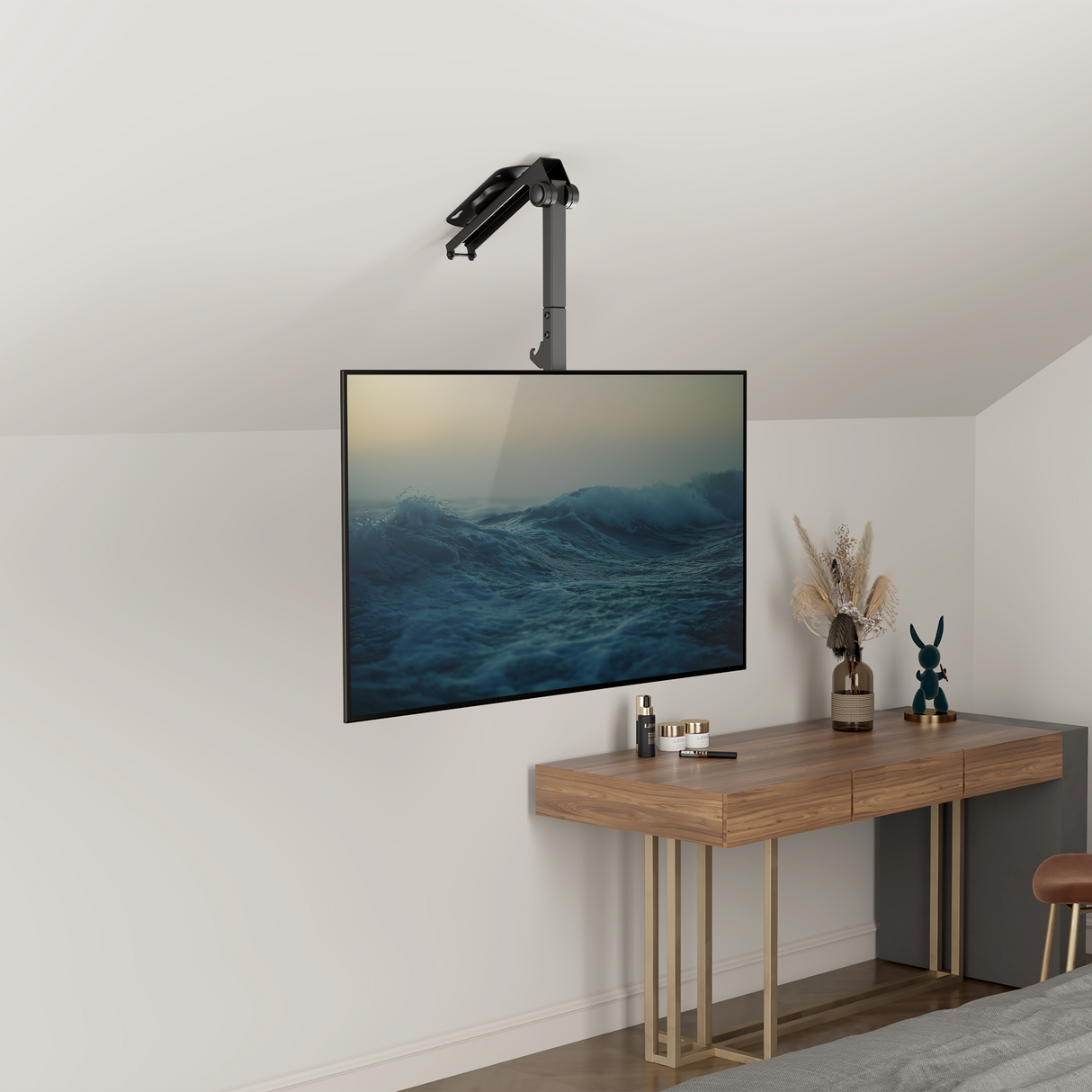 A flat-screen TV with the Mount-It! Flip-Down Ceiling TV Mount is installed above a wooden desk decorated with dried flowers, books, and a blue rabbit figurine in this modern minimalist room, displaying calming ocean waves.