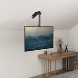 A flat-screen TV with the Mount-It! Flip-Down Ceiling TV Mount is installed above a wooden desk decorated with dried flowers, books, and a blue rabbit figurine in this modern minimalist room, displaying calming ocean waves.