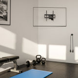 A home gym corner with a weight bench, dumbbells, kettlebells, a blue mat, and a jump rope on the wall. Above is an empty Mount-It! Full Motion Corner TV Wall Mount. Sunlight casts inviting shadows across the floor and walls.