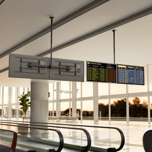 A modern airport departure area features a Mount-It! Dual TV Ceiling Mount with Long Pole holding two screens—one displaying flight info, the other blank—against glass walls, trees, and a cloudy sky beyond.
