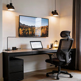 A modern home office with a black desk, ergonomic chair, laptop, desk lamp, lantern, and a wall-mounted photo of autumn trees and mountains. Two wall sconces add warm light alongside a Mount-It! Low-Profile Tilt TV Wall Mount.