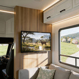 A modern RV interior has a TV displaying deer by a pond, mounted on the Mount-It! Low-Profile Tilt TV Wall Mount. Sunlight shines through a large window beside a gray sofa with a floral pillow. The space features light wood and white cabinets.