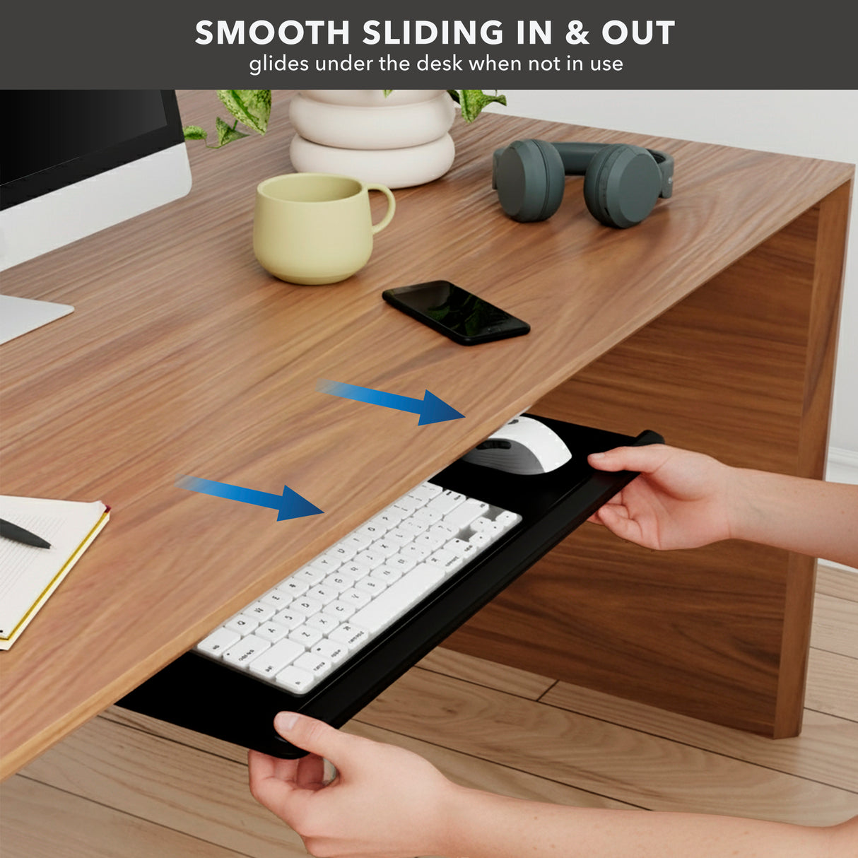 A person slides the Mount-It! Adjustable Keyboard Tray and Mouse Platform w/ Wrist Rest Pad under a wooden desk with accessories on top. Blue arrows show smooth sliding motion. Text above reads: “Smooth sliding in & out.”.