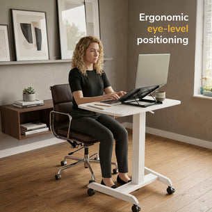 A woman works at a height-adjustable desk with her monitor on the Mount-It! Portable Folding Laptop Stand at eye level and a keyboard tray. Text reads, “Ergonomic eye-level positioning.” The scene is a modern home office with wall art and a window.