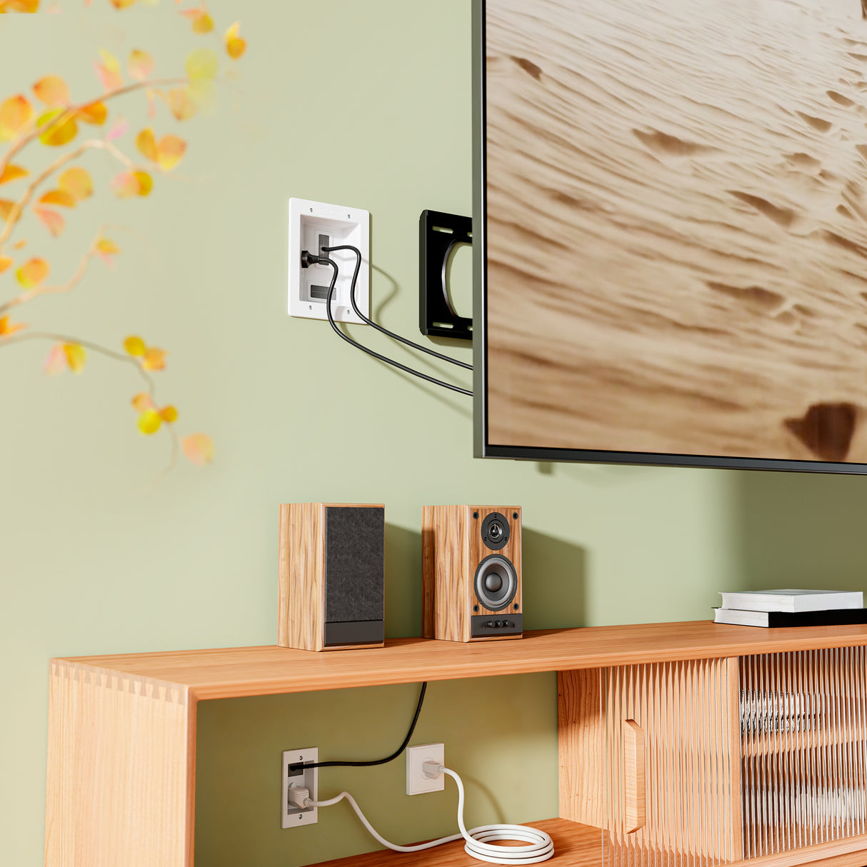 A wall-mounted TV with cables routed through the Mount-It! In-Wall Cable Management Kit with Power Input Module sits above a wooden media console holding speakers and books, while a leafy plant appears on the left side of the image.