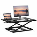 The Mount-It! Height Adjustable X-Lift Standing Desk Converter in black holds a monitor, laptop, keyboard, and mouse. Both screens show a sunset lake with mountains and trees, creating an ergonomic and flexible workspace setup.
