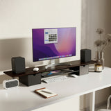 A modern white desk features the Mount-It! Extra Wide Monitor Riser with Drawers and Shelf, two black speakers, a wireless keyboard and mouse, a notebook with a pen, a candle, dried flowers in a vase, and neatly arranged office accessories.