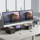 The Mount-It! Extra Wide Monitor Riser with Drawers and Shelf supports two monitors, plus a keyboard, smartphone, white mouse, water bottle, and pen—all neatly arranged in a bright office overlooking the city.