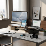 A bright, modern office features the Mount-It! Extra Wide Monitor Riser with Drawers and Shelf elevating a large screen, alongside a laptop, tablet, notebook, pen, mouse, keyboard, headphones, and art—all set against a city view.