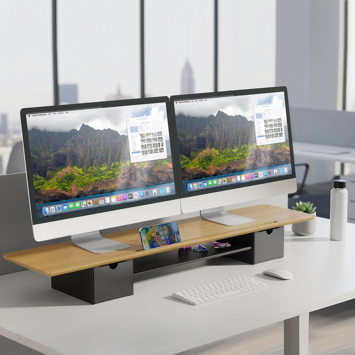 A Mount-It! Extra Wide Monitor Riser with Drawers and Shelf supports two monitors, a smartphone, wireless keyboard and mouse, and a water bottle, set in front of large windows overlooking a cityscape.
