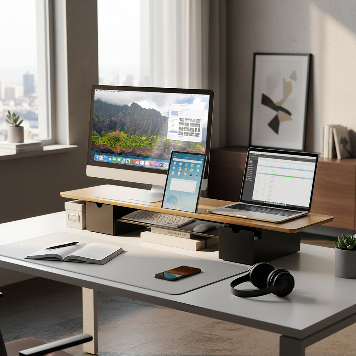 A minimalistic desk setup features the Mount-It! Extra Wide Monitor Riser with Drawers and Shelf, holding a desktop computer, laptop, tablet, notebook, smartphone, wireless headphones, and pen—all neatly organized with city views in the background.