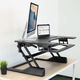 The Mount-It! Standing Desk Converter sits on a light wood desk, holding an iMac, laptop, and ergonomic keyboard tray. A small potted plant is on the left, set against a plain white wall in the background.