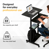 A man uses the Mount-It! Height Adjustable Rolling Stand up Desk with three black shelves for a laptop, books, and backpack. Locking casters provide stability. Each shelf supports 33 lbs; total capacity is 132 lbs—perfect for daily professional use.
