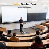 A teacher stands at a Mount-It! Height Adjustable Rolling Laptop Cart in front of a tiered classroom, addressing students who are seated and taking notes. Behind the teacher are large whiteboard and chalkboard surfaces.