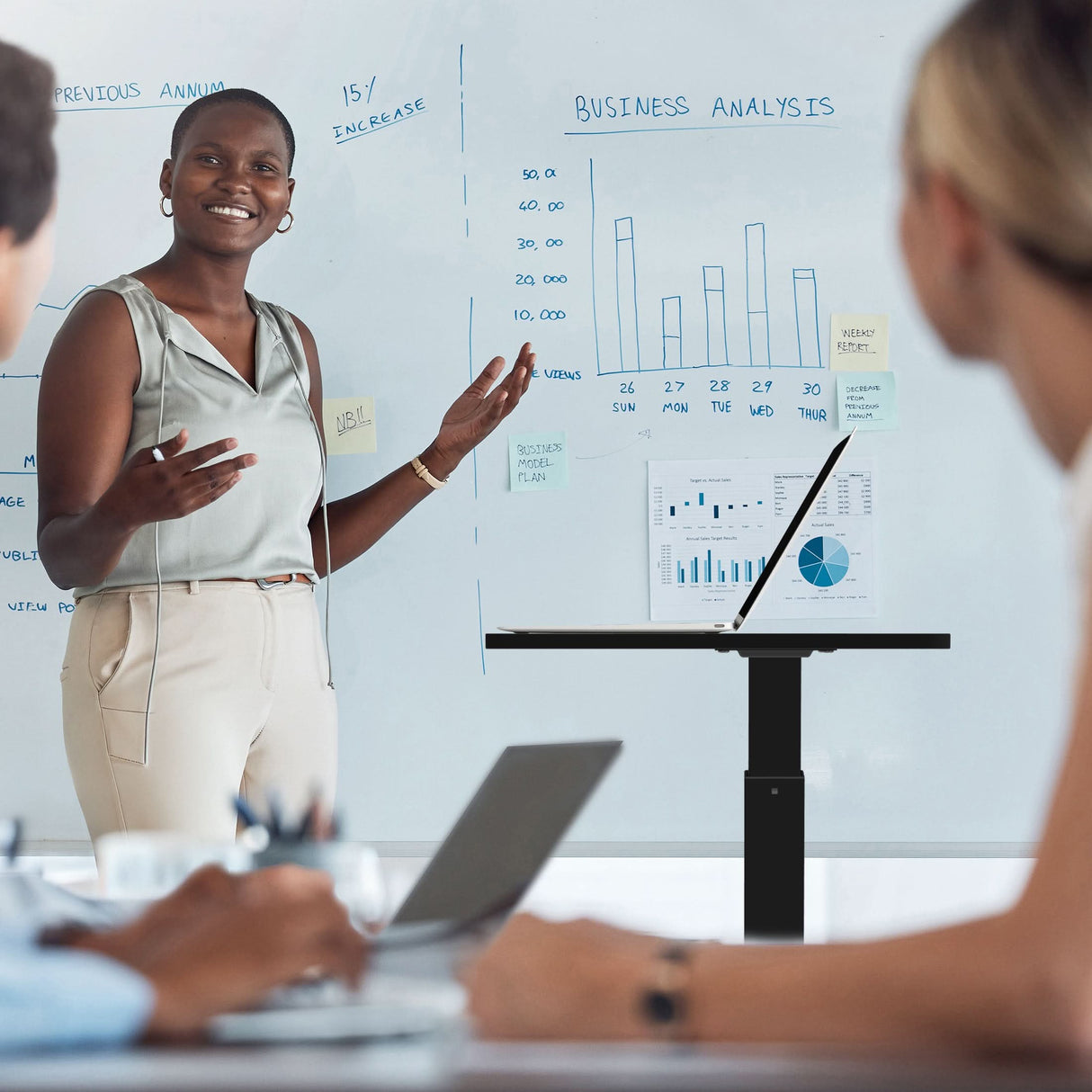 A woman presents data to two colleagues, using a Mount-It! Height Adjustable Rolling Laptop Desk for her laptop. The ergonomic workstation enhances comfort in this collaborative office, with charts and sticky notes on the whiteboard behind her.