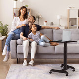 A smiling family of three sits on a gray sofa in a cozy living room, using the Mount-It! Height Adjustable Rolling Laptop Desk. The space features plants, a soft rug, and a light color scheme.