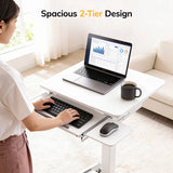 A woman uses the Mount-It! Laptop Cart with Pull-Out Keyboard Tray, a white mobile desk with a spacious 2-tier design and ergonomic tray for comfortable typing and device placement. A laptop, keyboard, mouse, and coffee mug sit on the desk.