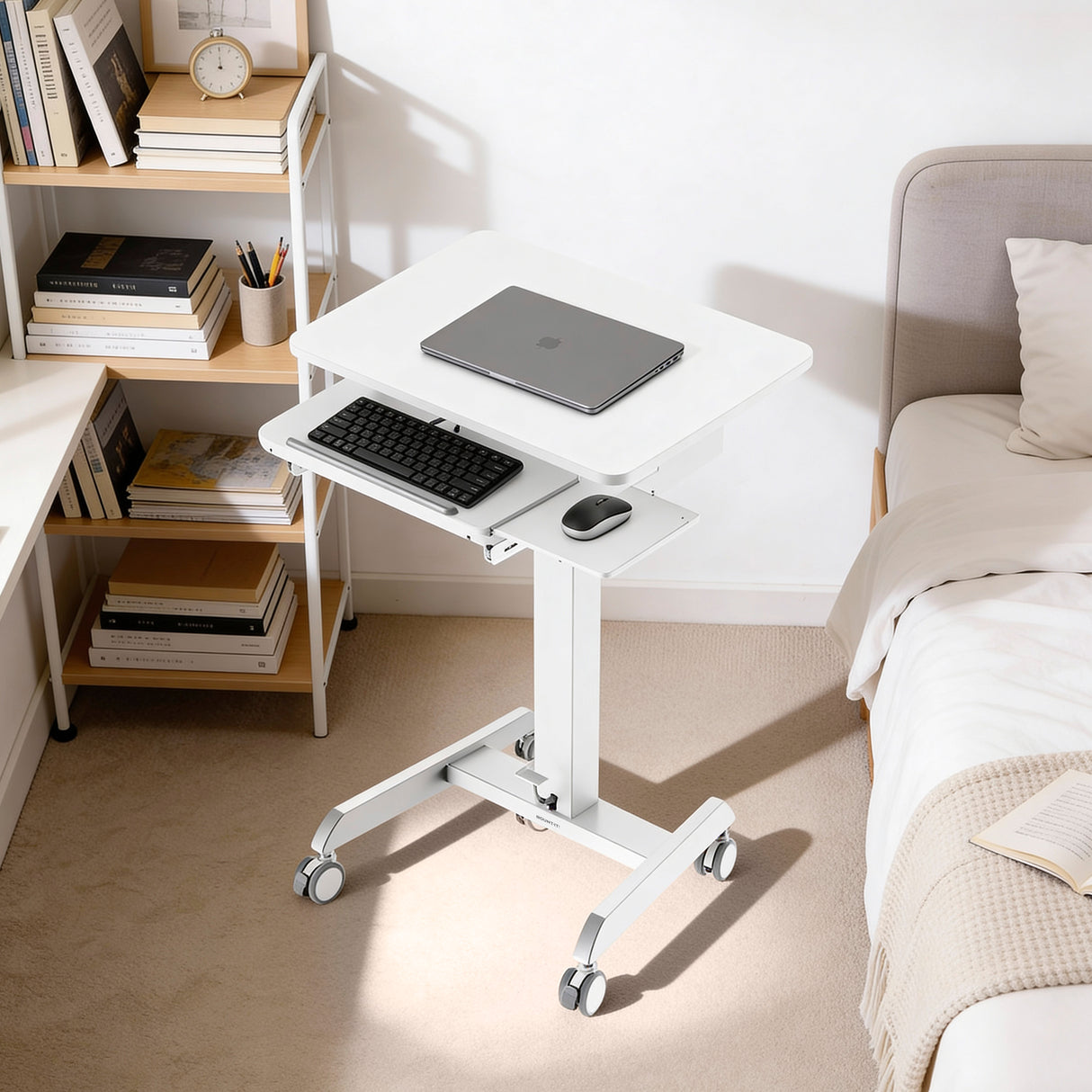 The Mount-It! Laptop Cart with Pull-Out Keyboard Tray, in white and adjustable in height, stands by a bed holding a laptop. Nearby, a bookshelf with books and a clock are softly illuminated by natural light.