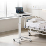 The Mount-It! Laptop Cart with Pull-Out Keyboard Tray, a height-adjustable and mobile standing desk, is positioned beside a hospital bed with folded towels in a bright, clean hospital room.