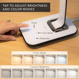 A hand taps the Turcom Relaxalight LED Desk Lamp by Mount-It! touch sensor base with labeled buttons for power, 5 color modes, and 7 brightness levels—each designed for eye protection. TAP TO ADJUST BRIGHTNESS AND COLOR MODES.