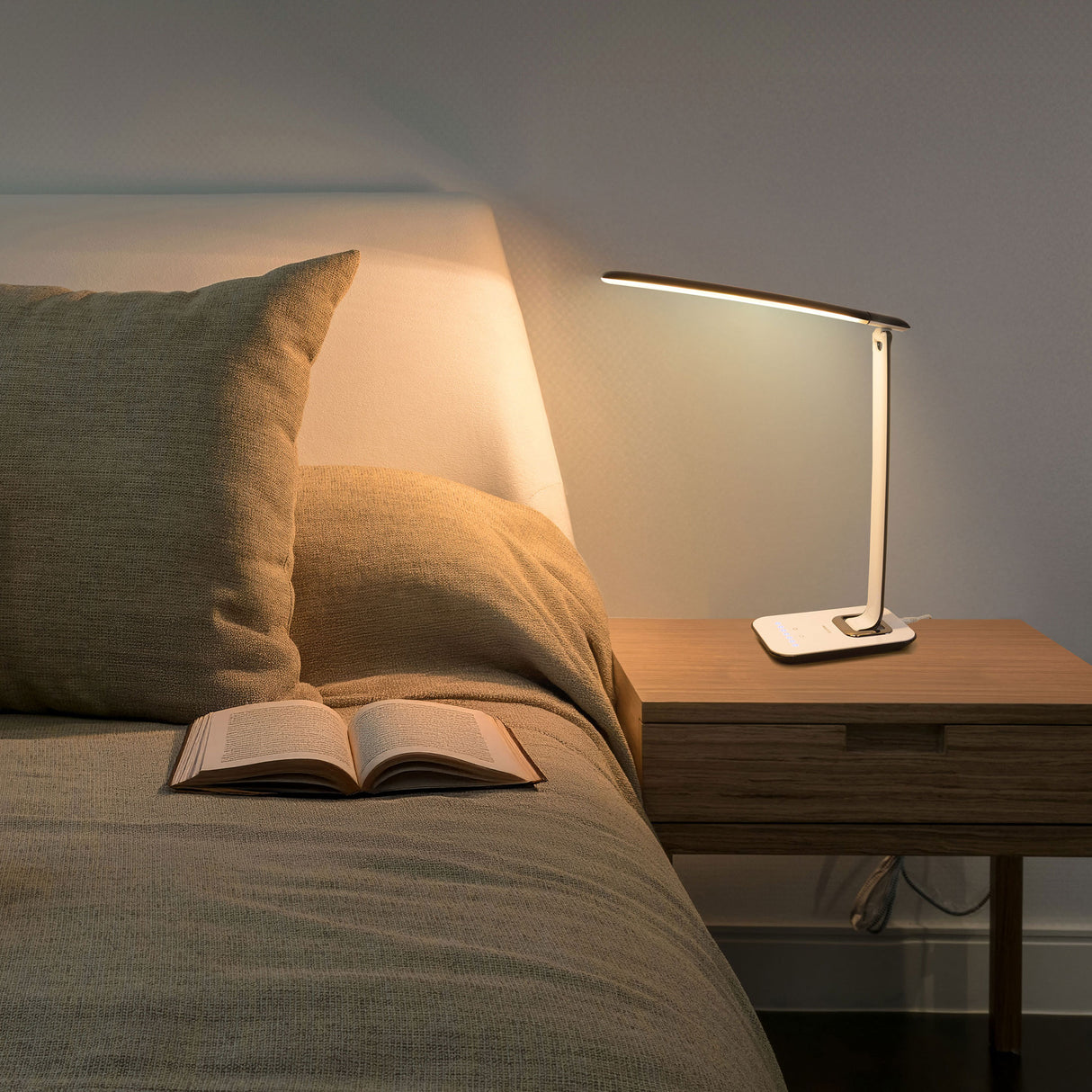 A neatly made bed with a beige pillow and blanket sits beside a wooden nightstand featuring the Mount-It! Turcom Relaxalight LED Desk Lamp with eye protection lighting turned on, and an open book lying on the bed.
