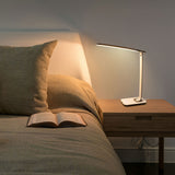 A neatly made bed with a beige pillow and blanket sits beside a wooden nightstand featuring the Mount-It! Turcom Relaxalight LED Desk Lamp with eye protection lighting turned on, and an open book lying on the bed.