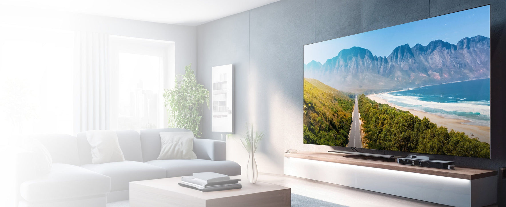A modern living room featuring a large flat-screen television mounted on a sleek white media console against a dark gray wall. The TV displays a scenic coastal road flanked by green trees and mountains under a blue sky. The room is bright with natural light coming from a large window on the left side, showcasing a comfortable white sectional sofa, a small coffee table with books, and a potted plant near the window.