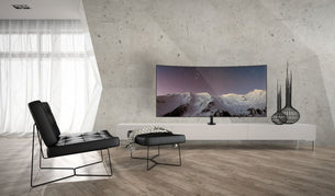 Modern minimalist living room with a black chair, ottoman, and a white console topped with a curved TV on the Mount-It! Bolt Down TV Stand. Decorative vases and sheer curtains finish the effortlessly stylish space.